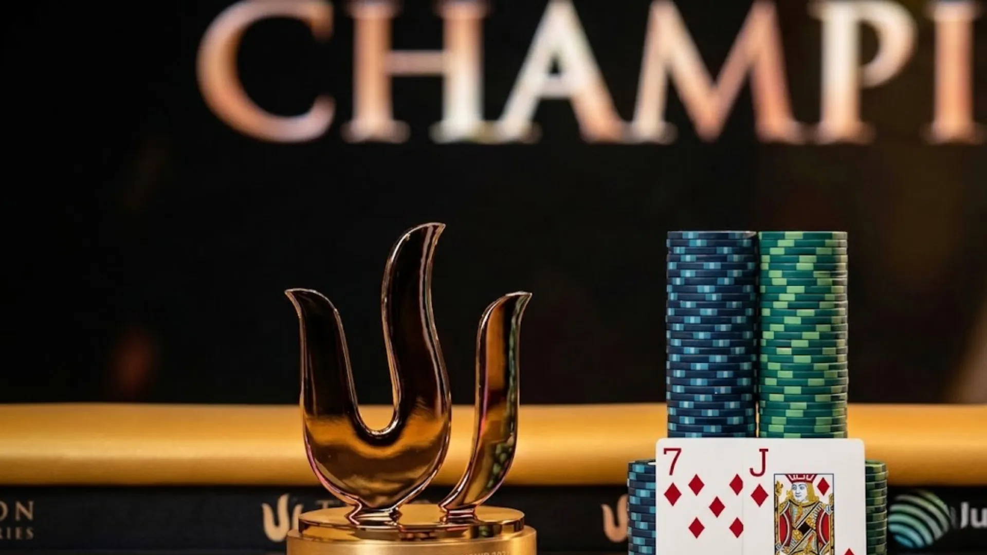 済州2026 Triton Pokerシリーズの金トロフィーとダイヤのJ-7勝利ハンドがチップスタックに立てかけられ、背景にCHAMPIONの文字